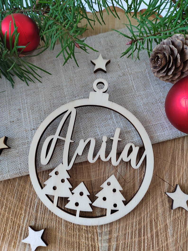 K&ouml;nnte beinhalten: Ein runder Weihnachtsbaumschmuck aus Holz mit dem ausgeschnittenen Namen "Anita" in der Mitte. Der Schmuck ist mit drei kleinen Weihnachtsb&auml;umen verziert und hat oben ein Loch zum Aufh&auml;ngen.