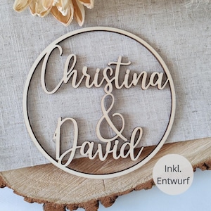 Könnte beinhalten: Ein kreisförmiges Holzschild mit den lasergeschnittenen Namen Christina & David in Schreibschrift. Das helle Holz hat dunkle, verbrannte Kanten vom Schneidevorgang. Ein kleiner runder Aufkleber mit dem Text Inkl. Entwurf ist ebenfalls zu sehen.