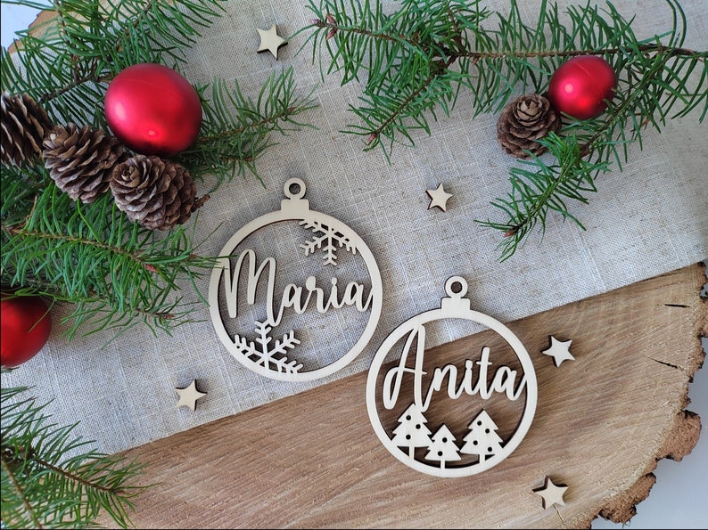 K&ouml;nnte beinhalten: Zwei h&ouml;lzerne Weihnachtsbaumschmuckst&uuml;cke mit den Namen "Maria" und "Anita" darauf. Das Ornament mit "Maria" hat Schneeflocken, und das Ornament mit "Anita" hat Weihnachtsb&auml;ume.