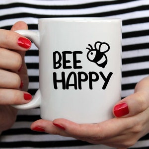 Puede incluir: Taza de cer&aacute;mica blanca con una ilustraci&oacute;n de abeja negra y el texto "bee happy".