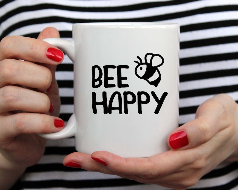 Puede incluir: Taza de cer&aacute;mica blanca con una ilustraci&oacute;n de abeja negra y el texto "bee happy".