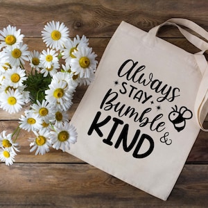 Puede incluir: Una bolsa de lona blanca con texto negro que dice "Always Stay Bumble & Kind" con una peque&ntilde;a ilustraci&oacute;n de abeja negra y amarilla. La bolsa est&aacute; rodeada de margaritas blancas sobre un fondo de madera.