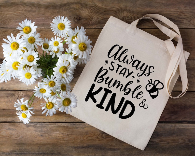 Puede incluir: Una bolsa de lona blanca con texto negro que dice "Always Stay Bumble & Kind" con una peque&ntilde;a ilustraci&oacute;n de abeja negra y amarilla. La bolsa est&aacute; rodeada de margaritas blancas sobre un fondo de madera.