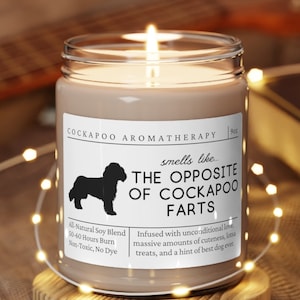 May include: A brown candle with a white label that reads "Cockapoo Aromatherapy" and "smells like... The Opposite of Cockapoo Farts". The label also includes the text "All Natural Soy Blend, 50-60 Hours Burn, Non-Toxic, No Dye" and "Infused with unconditional love, massive amounts of cuteness, treats, and a hint of best dog ever."