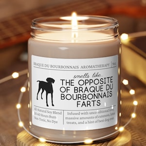 May include: A brown soy candle with a white label that reads "Braque du Bourbonnais Aromatherapy" and "smells like... The Opposite of Braque du Bourbonnais Farts". The label also includes a black and white illustration of a dog, and the text "All Natural Soy Blend, 50-60 Hours Burn, Non-Toxic, No Dye". The candle is sitting on a wooden coaster.