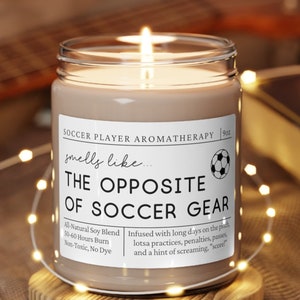 May include: A brown soy candle with a white label that reads "Soccer Player Aromatherapy" and "smells like... THE OPPOSITE OF SOCCER GEAR". The label also includes a soccer ball graphic and the text "All Natural Soy Blend, 50-60 Hours Burn, Non-Toxic, No Dye". The candle is sitting on a wooden coaster.