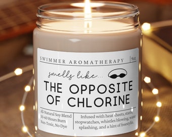 May include: A brown candle with a white label that reads "SWIMMER AROMATHERAPY" and "smells like... THE OPPOSITE OF CHLORINE". The label also includes details about the candle, such as "All Natural Soy Blend", "50-60 Hours Burn", and "Non-Toxic, No Dye". The candle is sitting on a wooden coaster.