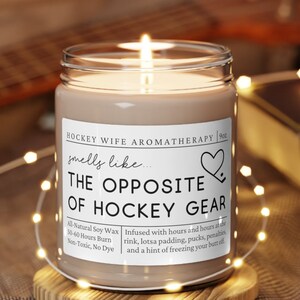 May include: A brown soy candle with a white label that reads "Hockey Wife Aromatherapy" and "smells like... THE OPPOSITE OF HOCKEY GEAR". The label also includes details about the candle, such as "All Natural Soy Wax", "50-60 Hours Burn", "Non-Toxic, No Dye", and "Infused with hours and hours at the rink, lotsa padding, pucks, penalties and a hint of freezing your best off."