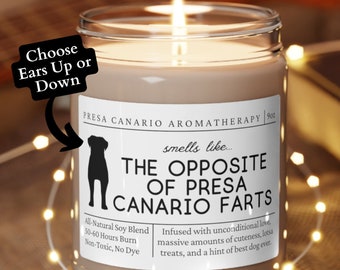 May include: A brown candle with a white label that reads "Presa Canario Aromatherapy" and "The Opposite of Presa Canario Farts". The candle is on a wooden coaster and surrounded by string lights.