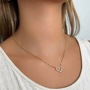 Op de afbeelding: Een gouden ketting met een delicaat hartvormig hanger. De ketting wordt gedragen door een persoon met lang bruin haar.