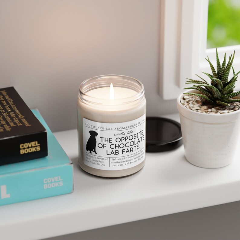 Może przedstawiać: Biała świeca z czarną sylwetką psa na etykiecie. Etykieta zawiera napis "Chocolate Lab Aromatherapy" i "The Opposite of Chocolate Lab Farts".