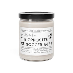 May include: A white candle jar with a black lid and a soccer ball graphic. The label reads "Soccer Player Aromatherapy" and "smells like... THE OPPOSITE OF SOCCER GEAR". The candle is infused with a scent inspired by long days on the pitch, practices, penalties, passes, and a hint of screaming. "Score!" The candle is made with an all-natural soy blend and has a 50-60 hour burn time. It is non-toxic and dye-free.