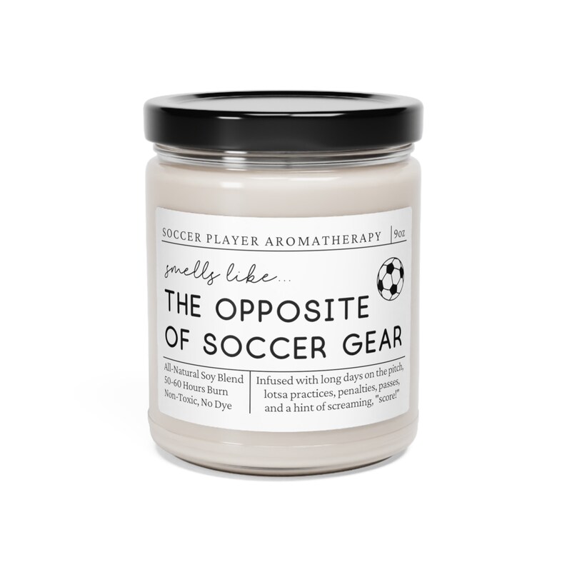 May include: A white candle jar with a black lid and a soccer ball graphic. The label reads "Soccer Player Aromatherapy" and "smells like... THE OPPOSITE OF SOCCER GEAR". The candle is infused with a scent inspired by long days on the pitch, practices, penalties, passes, and a hint of screaming. "Score!" The candle is made with an all-natural soy blend and has a 50-60 hour burn time. It is non-toxic and dye-free.