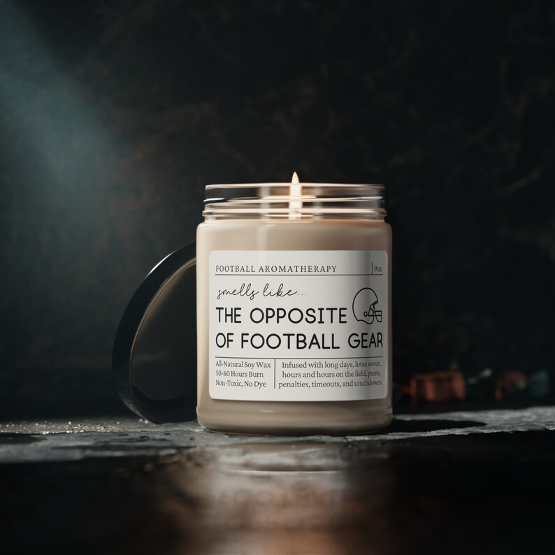Może przedstawiać: Beżowa świeca z czarną etykietą, na kt&oacute;rej widnieje napis "Football Aromatherapy smells like... THE OPPOSITE OF FOOTBALL GEAR". Etykieta zawiera r&oacute;wnież informacje o świecy, takie jak "All-Natural Soy Wax", "50-60 Hours Burn", "Non-Toxic, No Dye" oraz "Infused with long days, lotsa sweat, hours and hours on the field, penalties, timeouts, and touchdowns".