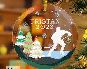 May include: A round glass Christmas ornament with a hockey player silhouette, snowflakes, and trees. The ornament is decorated with a brown, orange, and yellow gradient background. The text "TRISTAN 2023" is printed on the ornament.