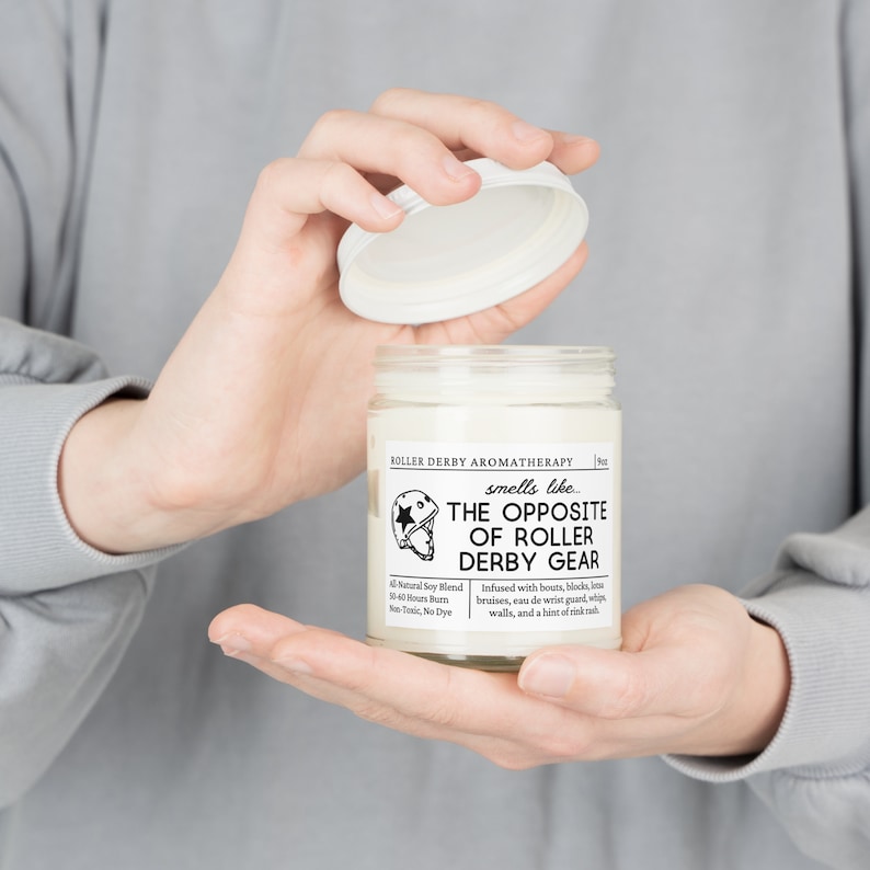 Może przedstawiać: Biały słoik ze świecą z czarną i białą etykietą, na kt&oacute;rej widnieje napis "Roller Derby Aromatherapy pachnie jak... Przeciwieństwo sprzętu do roller derby". Etykieta zawiera r&oacute;wnież małą ilustrację kasku do roller derby. Słoik ze świecą jest trzymany przez dłonie osoby.