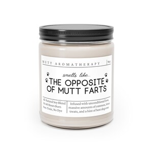 May include: A beige candle jar with a black lid and a white label that reads "MUTT AROMATHERAPY" and "smells like... THE OPPOSITE OF MUTT FARTS". The label also includes the text "All-Natural Soy Blend, 50-60 Hours Burn, Non-Toxic, No Dye" and "Infused with unconditional love, massive amounts of cuteness, lotsa treats, and a hint of best dog ever."
