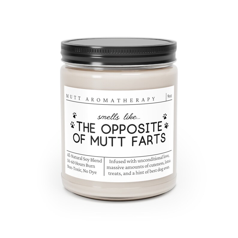 May include: A beige candle jar with a black lid and a white label that reads "MUTT AROMATHERAPY" and "smells like... THE OPPOSITE OF MUTT FARTS". The label also includes the text "All-Natural Soy Blend, 50-60 Hours Burn, Non-Toxic, No Dye" and "Infused with unconditional love, massive amounts of cuteness, lotsa treats, and a hint of best dog ever."