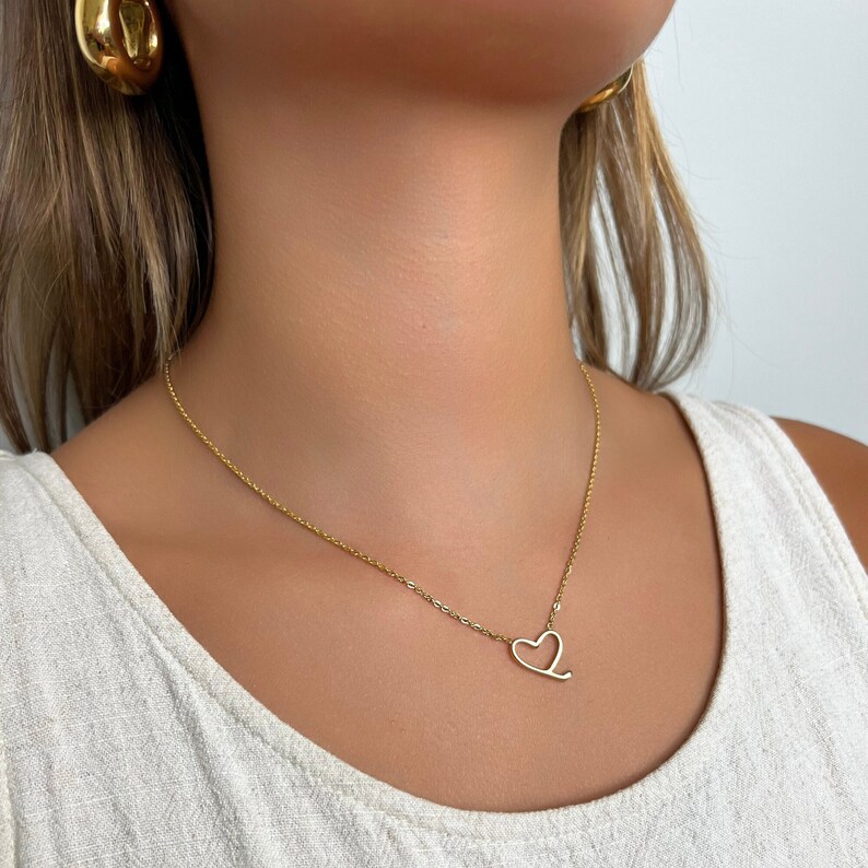 Op de afbeelding: Een gouden ketting met een delicaat hartvormig hanger. De ketting wordt gedragen door een persoon met lang bruin haar.