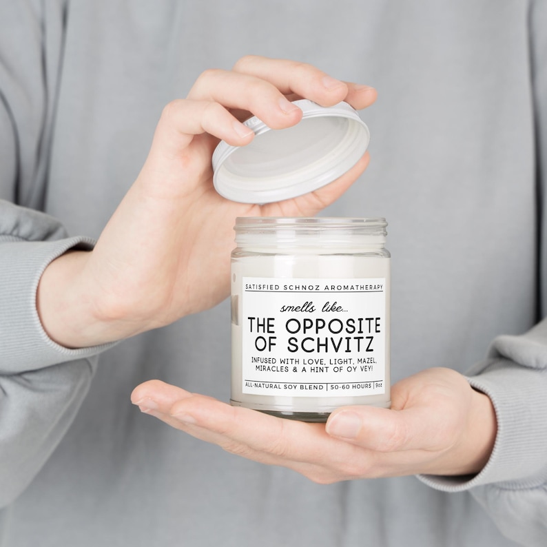 May include: A white candle jar with a black label that reads "SATISFIED SCHNOZ AROMATHERAPY" and "smells like... THE OPPOSITE OF SCHVITZ"  "INFUSED WITH LOVE, LIGHT, MAZEL MIRACLES & A HINT OF OY VEY"  "ALL NATURAL SOY BLEND | 50-60 HOURS"