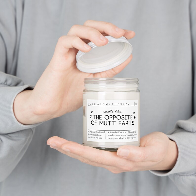 May include: A white candle jar with a black label that reads "MUTT AROMATHERAPY smells like... *THE OPPOSITE* OF MUTT FARTS". The label also includes details about the candle, such as the soy blend, burn time, and fragrance.