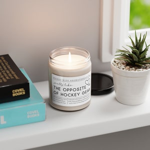 May include: A white candle with a brown label that reads "Hockey Wife Aromatherapy Jar" and "smells like... The Opposite of Hockey Gear". The candle is sitting on a white surface with two books and a potted succulent plant.
