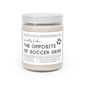 May include: A beige candle jar with a silver lid. The label reads "Soccer Player Aromatherapy" and "smells like... THE OPPOSITE OF SOCCER GEAR". The candle is infused with a blend of scents inspired by soccer, including long days on the pitch, practices, penalties, passes, and a hint of screaming "score".