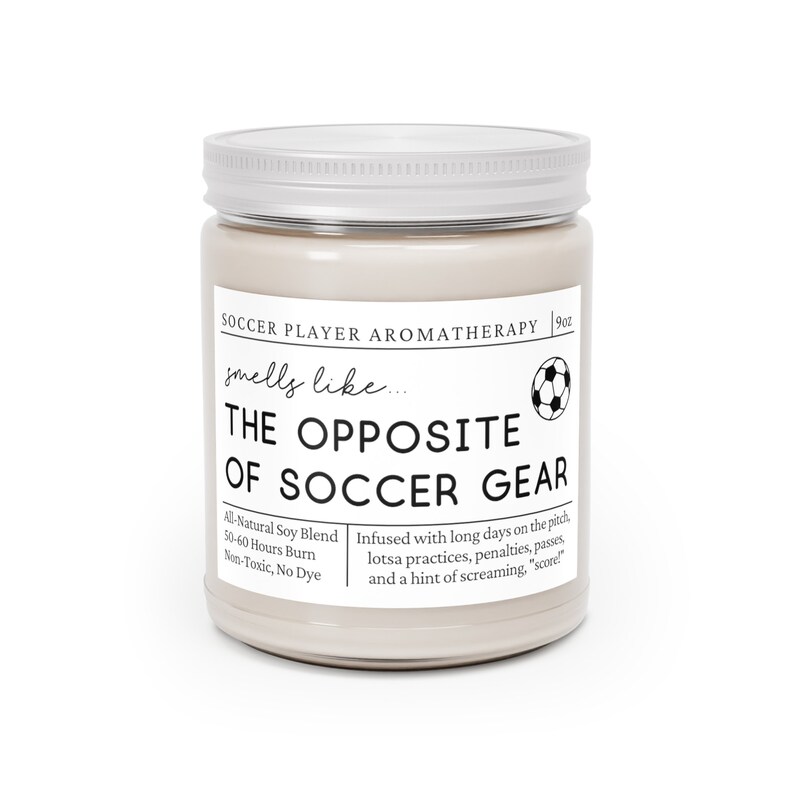 May include: A beige candle jar with a silver lid. The label reads "Soccer Player Aromatherapy" and "smells like... THE OPPOSITE OF SOCCER GEAR". The candle is infused with a blend of scents inspired by soccer, including long days on the pitch, practices, penalties, passes, and a hint of screaming "score".