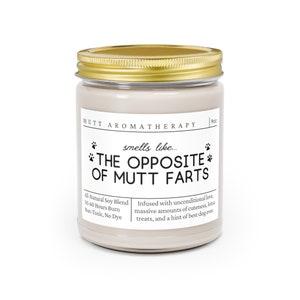 May include: A white candle jar with a gold lid. The label reads "Mutt Aromatherapy" and "smells like... THE OPPOSITE OF MUTT FARTS". The candle is infused with unconditional love, cuteness, treats, and a hint of best dog ever. It is made with a natural soy blend and burns for 50-60 hours.