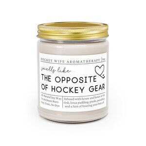 May include: A 9oz candle in a glass jar with a gold lid. The label reads "HOCKEY WIFE AROMATHERAPY" and "smells like... THE OPPOSITE OF HOCKEY GEAR". The candle is infused with scents of a hockey rink, padding, pucks, penalties, and a hint of freezing your butt off. The candle is made with all-natural soy wax and has a 50-60 hour burn time. It is non-toxic and dye-free.