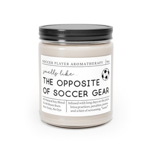 May include: A beige candle jar with a black lid and a white label that reads "Soccer Player Aromatherapy 9 oz. smells like... THE OPPOSITE OF SOCCER GEAR Al-Natural Soy Blend 50-60 Hours Burn Non-Toxic, No Dye Infused with long days on the pitch, lotsa practices, penalties, passes and a hint of screaming, "score!" "