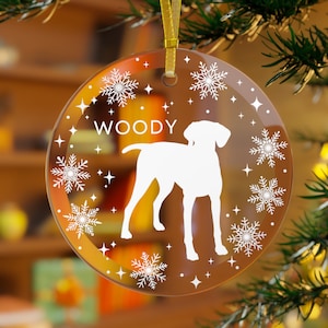May include: A clear glass Christmas ornament with a white silhouette of a dog and the name "WOODY" printed on it. The ornament is decorated with snowflakes and stars.