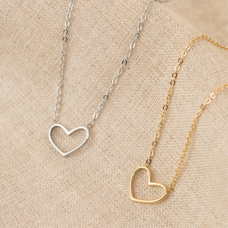May include: Two delicate necklaces with open heart pendants. One necklace is silver and the other is gold. The necklaces are displayed on a beige fabric background.