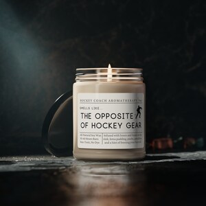 May include: A beige candle with a black lid. The candle is labeled "Hockey Coach Aromatherapy" and "The Opposite of Hockey Gear". The label also includes details about the candle, such as the type of wax, burn time, and scent.