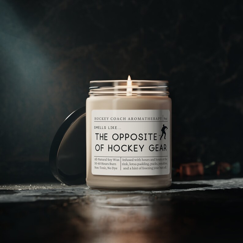 May include: A beige candle with a black lid. The candle is labeled "Hockey Coach Aromatherapy" and "The Opposite of Hockey Gear". The label also includes details about the candle, such as the type of wax, burn time, and scent.