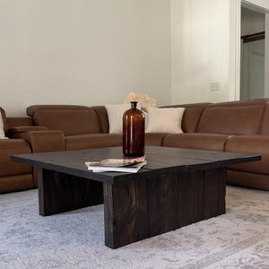 May include: A dark stained, square coffee table with a thick, solid top and a matching base. A brown glass vase with dried flowers sits on the table, along with a magazine. The table is in a living room with a brown leather sectional sofa.