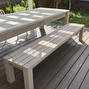 May include: A light-colored wooden outdoor dining set, including a rectangular table and a matching bench. The table and bench are constructed with a slatted design and are set on a wooden deck. The scene is bathed in sunlight, creating shadows.