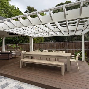 Puede incluir: Patio al aire libre con una pérgola blanca, una gran mesa de madera y un banco. La terraza es de material compuesto marrón. Se ven un brasero negro y una zona de estar.