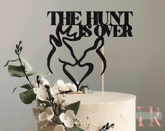 May include: Black glitter cake topper with the text "THE HUNT IS OVER" and two deer antlers forming a heart.