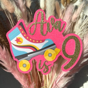 Pode incluir: Topper de bolo de glitter rosa e dourado com um design de patins e o texto "Ava faz 9 anos".