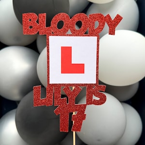 以下が含まれることがあります： 赤いグリッターのケーキトッパーに「Bloody Lily is 17」の文字と、赤い「L」が書かれた白い正方形があります。トッパーは白い風船に囲まれています。