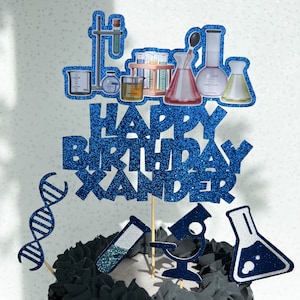 May include: Blue glitter cake topper with the words "Happy Birthday Xander" and science themed decorations including beakers, test tubes, and a microscope.