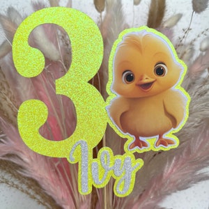 Op de afbeelding: Een geel glittercijfer 3 met een gele kuikenillustratie en het woord "1e" in zilveren glitter. De achtergrond is een boeket gedroogde bloemen.