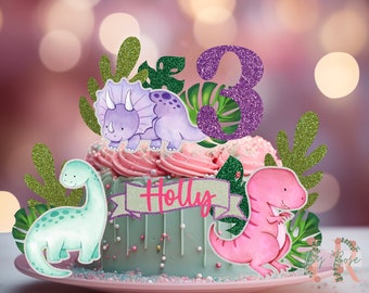 May include: A green, pink, and purple cake topper with a dinosaur theme. The topper includes a purple dinosaur, a pink dinosaur, green leaves, and the number 3. The cake topper also includes a banner with the name "Holly" on it.