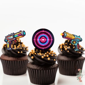 Könnte beinhalten: Drei Cupcakes mit schwarzem Zuckerguss und goldenen Streuseln. Jeder Cupcake ist mit einem anderen Laserpistolen- oder Zielscheiben-Design verziert. Die Designs sind farbenfroh mit Neon-Akzenten, ideal für eine Videospiel-Mottoparty.