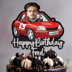 May include: A cake topper featuring a red car with a person driving, the word "LEARNER" on the car, and the text "Happy Birthday Fred" in silver glitter. The cake is decorated with chocolate and cream-colored confections.