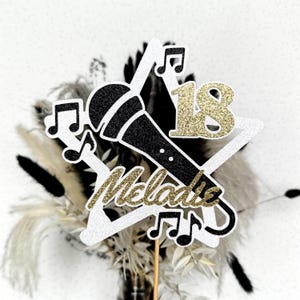 Puede incluir: Topper de pastel en forma de estrella blanca con un micrófono negro, '18' en purpurina dorada y el nombre 'Melodic' en purpurina dorada. El topper está decorado con notas musicales.
