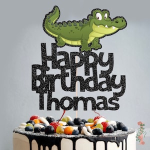 May include: A black glitter cake topper with the words "Happy Birthday Thomas" and a green alligator cartoon.
