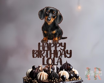 May include: A dachshund cake topper with the words "HAPPY BIRTHDAY LOLA" in brown glitter. The dog is black and brown, perched above a decorated cake with chocolate and white accents. A festive decoration for a birthday celebration.