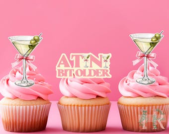 May include: Three pink cupcakes with white frosting. Each cupcake has a martini glass topper with a green olive. The middle cupcake has a pink and white sign that says "A TIN BIT OLDER".